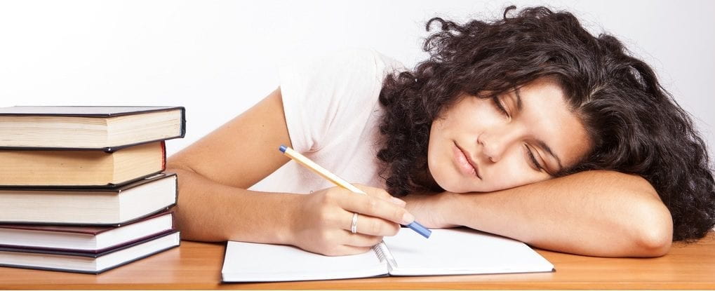 A woman writing with a pencil in a book. She is resting on her arm in a sleeping position.
